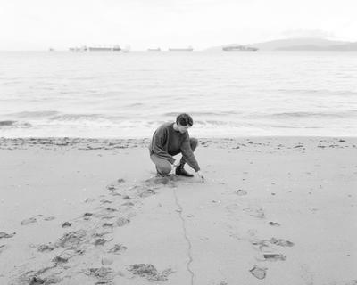 Person squats to touch the sand on the shore of a beach. Black and white.
