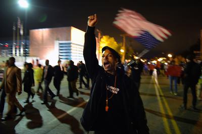 A male protestor yelling in front of a crowd of other protestors, one of his hands raised in a fist and the other holding an American flag