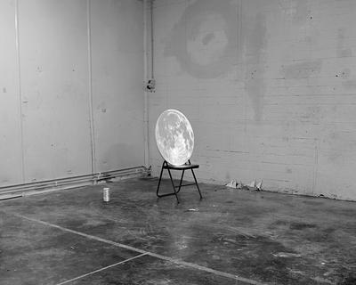 A folding chair sitting in an empty industrial room, an image of the moon sitting in the chair