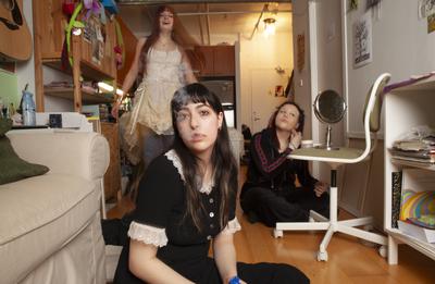 Two girls sit on the floor, one jumps up behind them. The girl closest to the camera is exhaling smoke. The one behind her surveys herself in a cosmetic mirror, propped on an office chair.