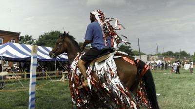 A Black man rides away from the camera on a horse decorated in silver and red streamers.