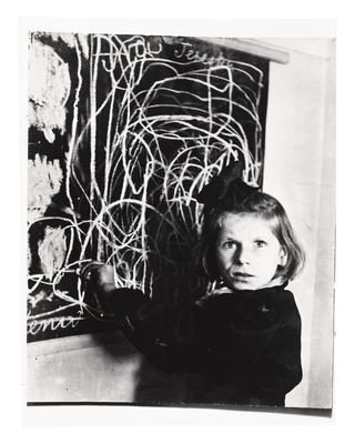 Little girl with short haircut and arm outstretched to scribble on chalkboard.