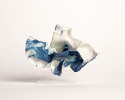 A flowing piece of fabric in indigo blue and white, resembling waves, is elegantly draped on a clear stand against a plain white background.