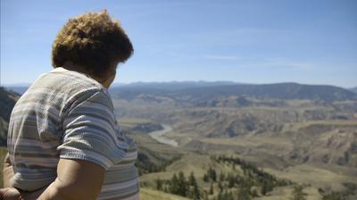 A woman wearing a striped shirt looks out over a landscape, her hands behind her back