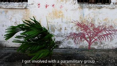 A still from a film. A plant blowing in the wind in front of an old building. Closed captioning reads: "I got involved with a paramilitary group."